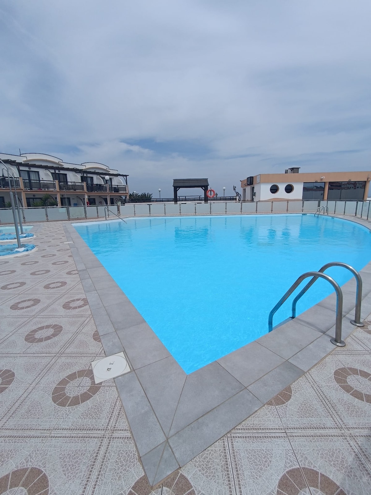 A large swimming pool is set within a tiled area, featuring a light blue hue. A gentle slope leads to the water, with a railing for support. Surrounding the pool are lounge chairs and a few shaded seating areas, enhancing accessibility for relaxation.