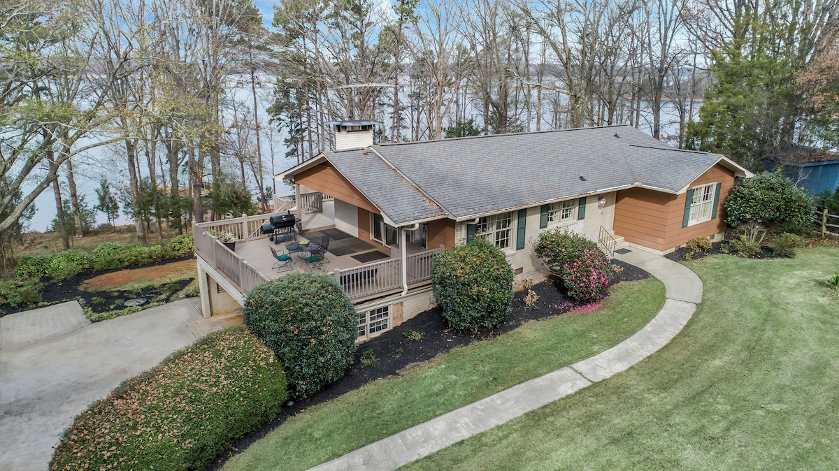 The exterior of the house is displayed with a clear view of the surrounding landscape. A spacious deck is shown with seating, and lush greenery is present along the pathway and garden beds. Nearby trees and a tranquil body of water contribute to the peaceful setting.