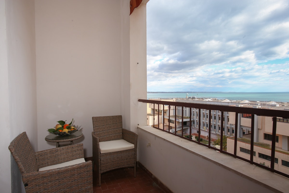 A balcony area is displayed, furnished with two woven chairs beside a small glass table. A bowl of fruit is present on the table, and the sea can be seen in the distance, with cloudy skies above. The railing offers an unobstructed view of the surroundings.