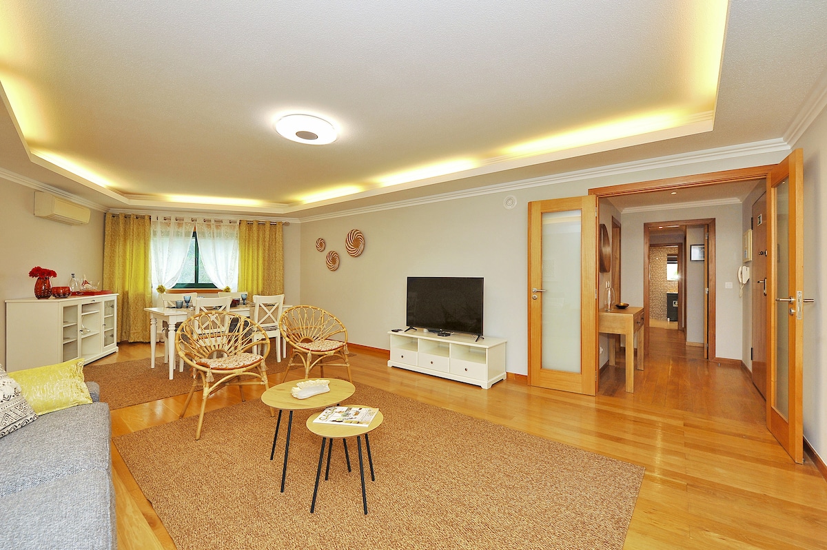 A spacious living area features warm wood flooring and subtle recessed lighting. A cozy sofa is positioned alongside decorative chairs, while a flat-screen TV is placed on a white media unit. A dining table is visible in the background, complemented by natural light filtering through the curtains.