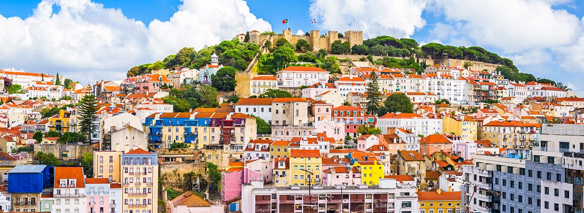 A panoramic view of a hillside cityscape showcases buildings with varying colors and rooftops, predominantly in reds and whites. The backdrop features a historic castle and lush greenery at the summit, under a bright sky adorned with scattered clouds.