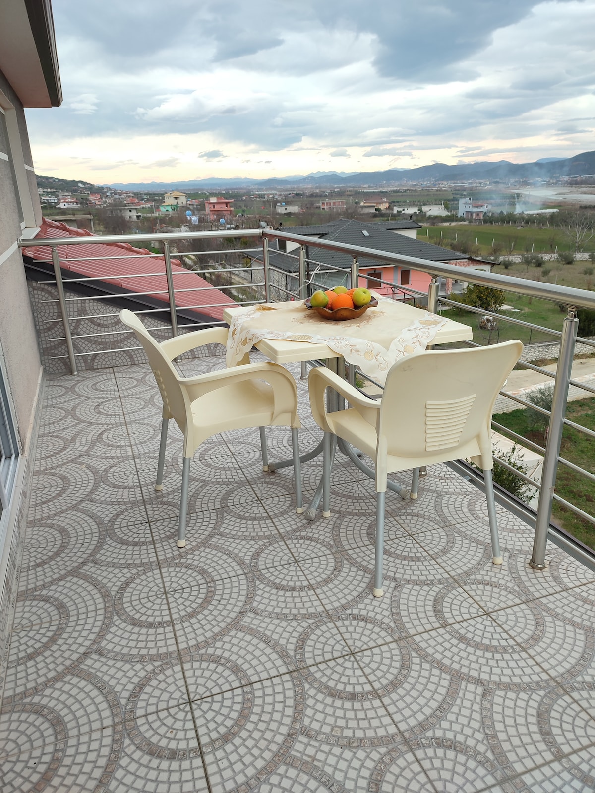 A balcony is visible, featuring a small table set with a bowl of fruit and two chairs. The floor has a circular patterned tile design. In the background, a scenic view of mountains and village rooftops can be seen under a cloudy sky.