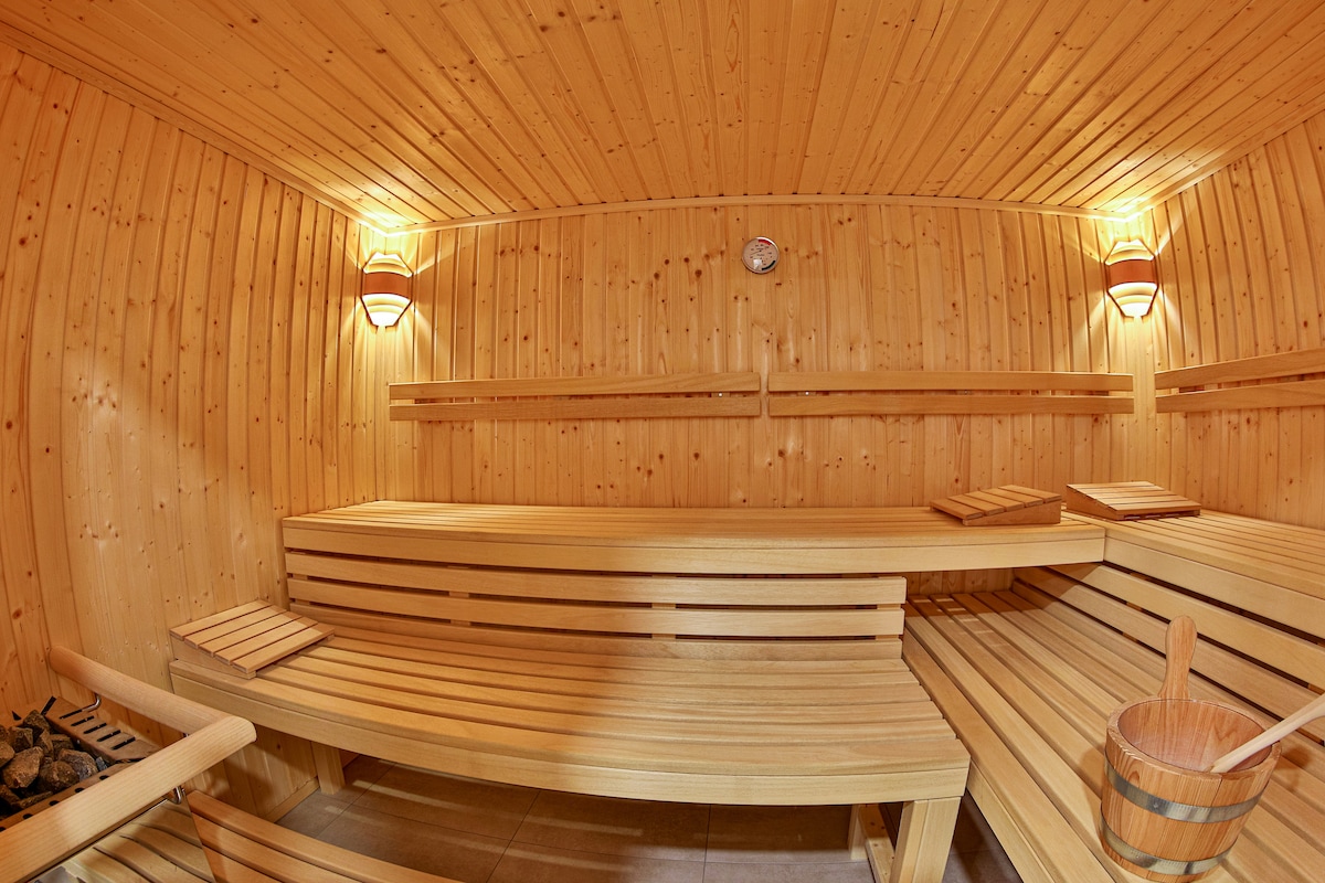 A wooden sauna features warm-toned wood paneling and benches arranged around the space. Wall sconces provide soft lighting, while a clock is mounted on one wall. A wooden bucket and ladle are positioned nearby, complementing the tranquil, relaxing atmosphere.
