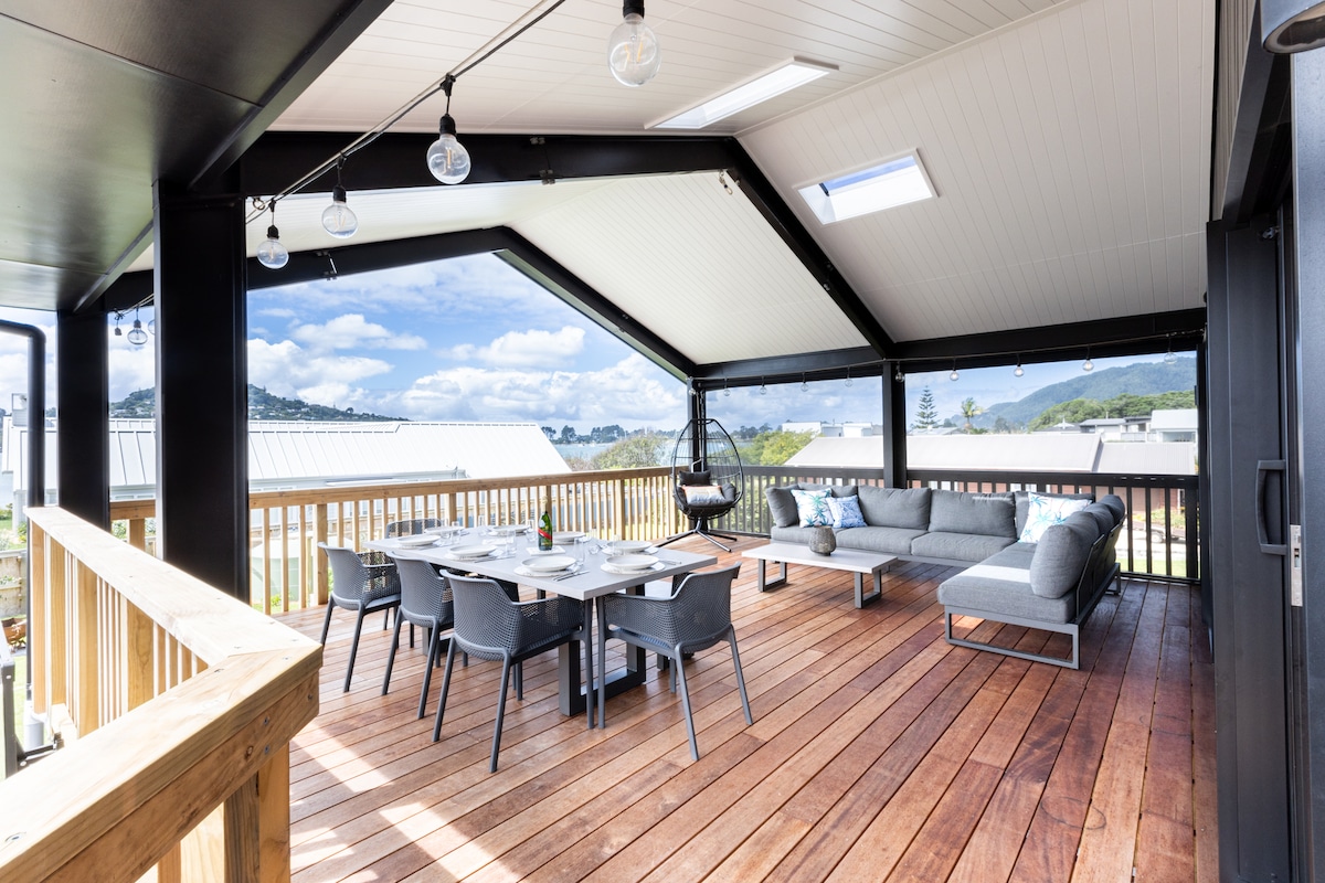 An outdoor covered deck features a spacious seating area with a sectional sofa and a dining table set. Clear views of the surrounding landscape are provided by large openings, enhancing the relaxed atmosphere. A ceiling fan and string lights add to the comfort of the space.