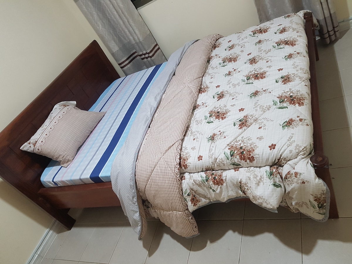 A cozy sleeping area features a wooden bed with a striped blue and white mattress. Soft floral-patterned bedding drapes over the spacious bed, complemented by a matching pillow. Natural light filters through partially opened curtains, illuminating the room's neutral color scheme.