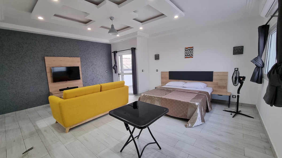 A spacious studio features a yellow sofa, a large bed with neutral bedding, and a sleek black table. A flat-screen TV is mounted on a wooden panel, and large windows provide natural light, complemented by dark curtains. The room is accented with modern decor elements.