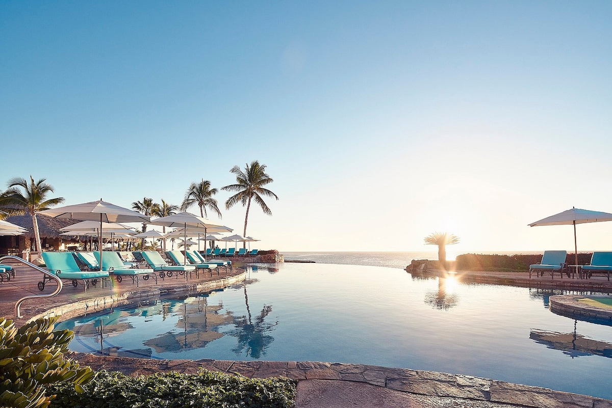 An inviting pool area is surrounded by comfortable lounge chairs and umbrellas, offering a picturesque view of the ocean. Palm trees sway gently in the breeze, while the sun rises, casting warm light across the tranquil water.