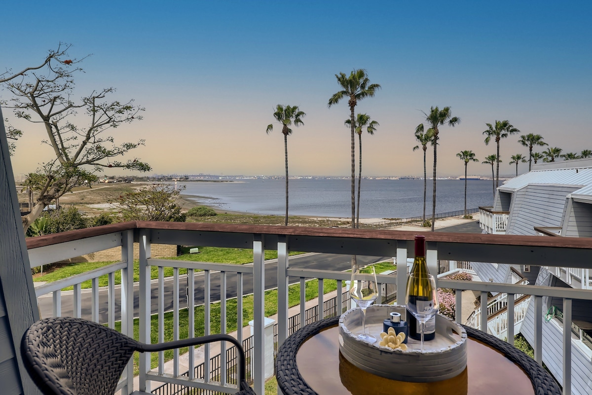 A balcony offers a scenic view of the bay, framed by palm trees. A small table is set with a bottle of wine and snacks, inviting relaxation while overlooking the tranquil water and distant city skyline.
