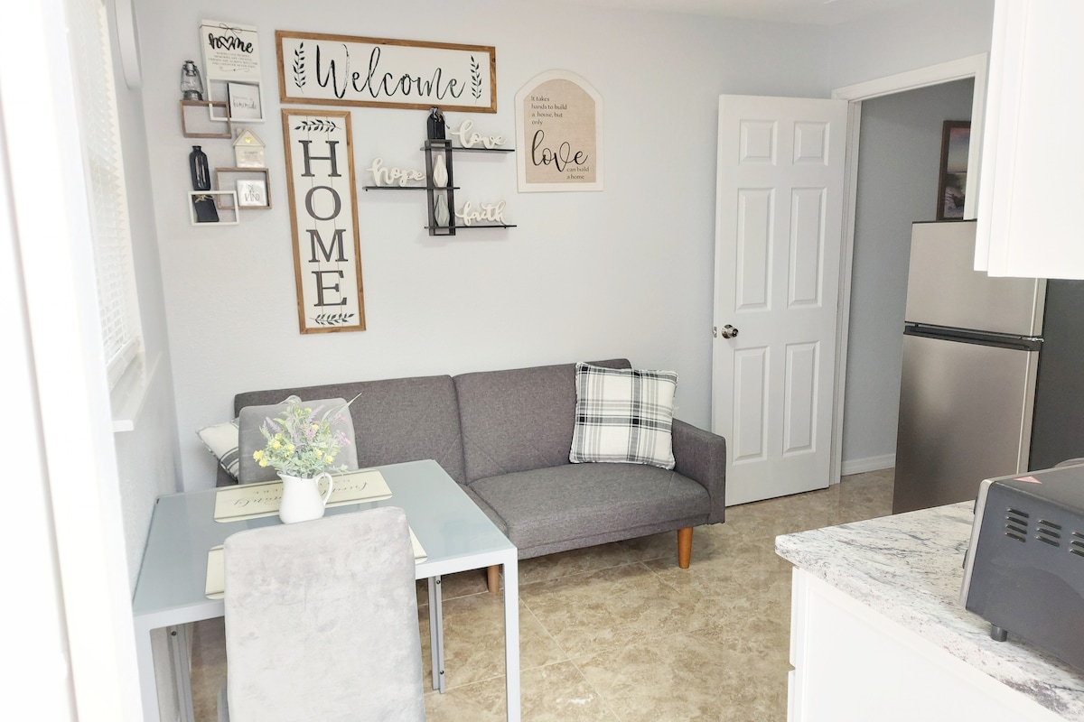 A compact dining area features a gray couch paired with a glass-top table. Decorative wall signs convey messages of welcome and love. A small floral arrangement sits on the table, while a refrigerator is visible in the background, completing the open and inviting space.
