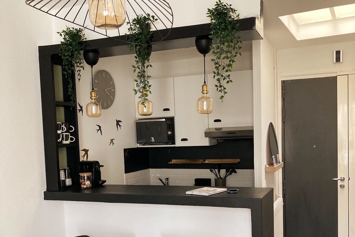 A modern kitchenette is visible, featuring a sleek black countertop with hanging plants. Three decorative pendant lights illuminate the space, complemented by a wall clock and a stylish coffee maker. A microwave and a stove are integrated into the cabinetry, enhancing the functional design.