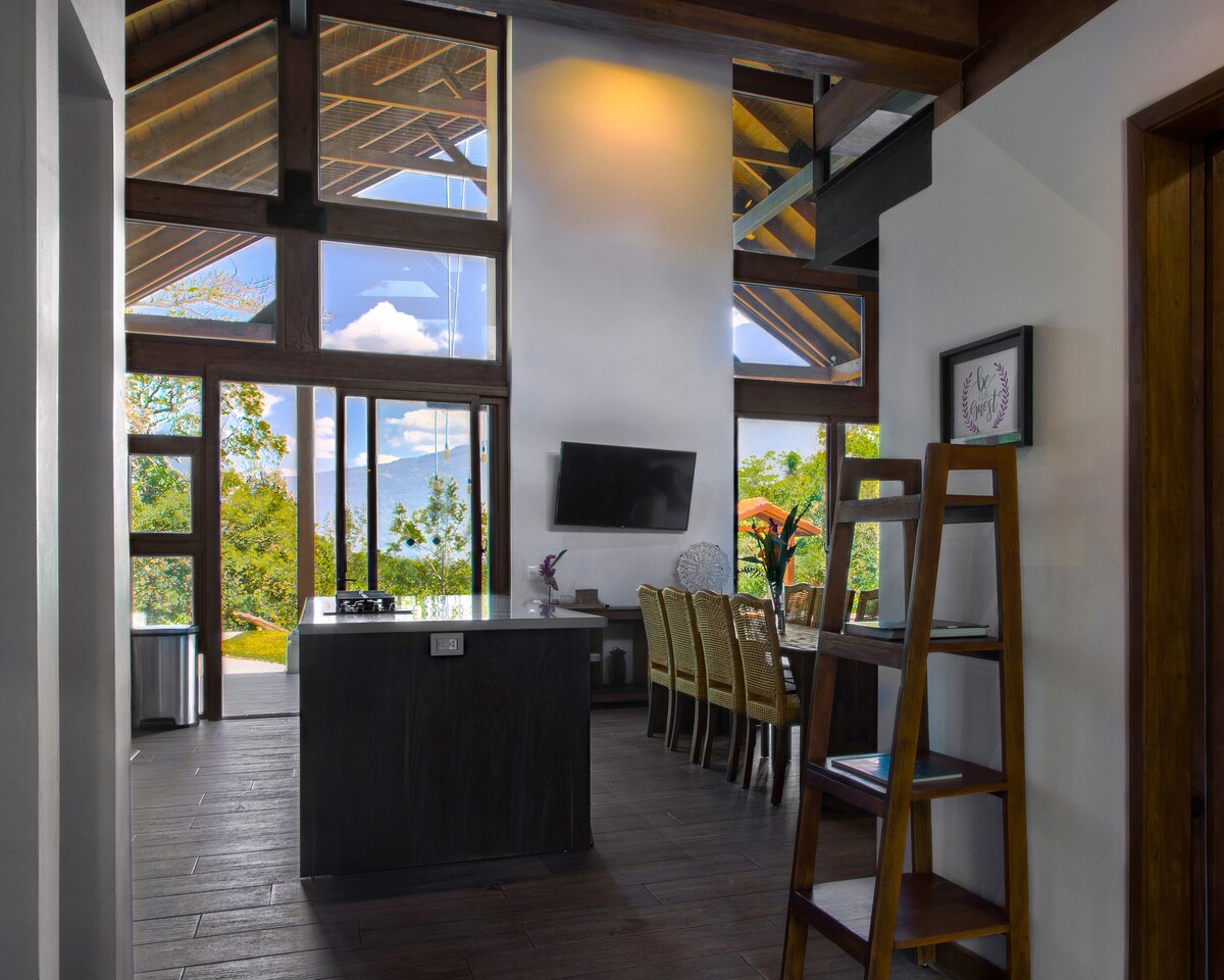 An open living area features high ceilings and large windows, providing views of the surrounding nature. A dining table accommodates multiple chairs, and a modern television mounted on the wall complements the space. Natural light pours in, creating a warm ambiance.