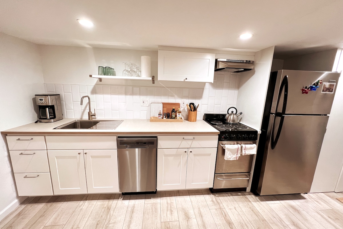 A modern kitchen is equipped with white cabinetry and stainless-steel appliances, including a dishwasher, oven, and refrigerator. A double bowl sink sits below a wall-mounted shelf, with kitchen tools arranged neatly. Ample counter space is available for meal preparation.