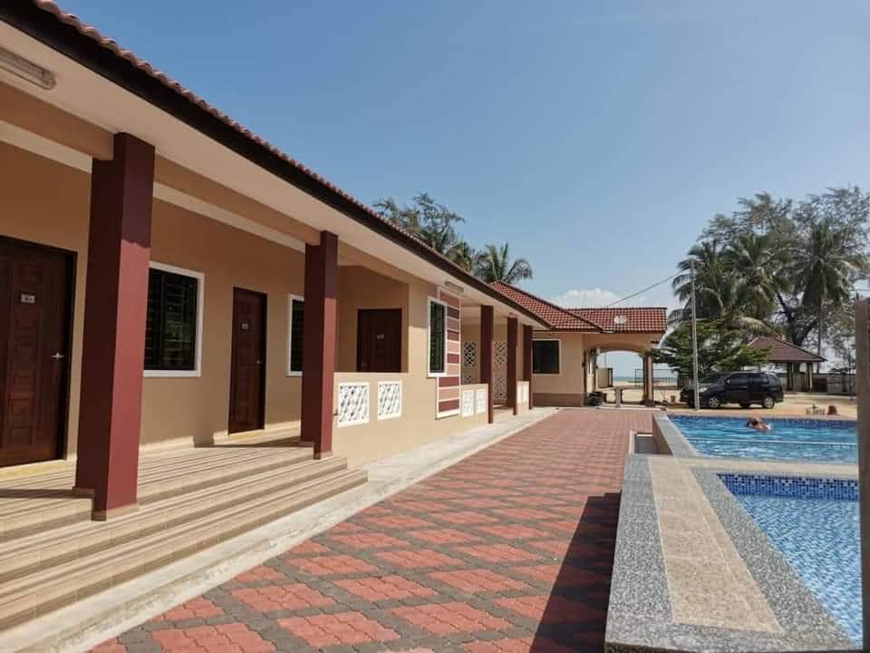 A well-maintained walkway is lined with several doorways leading to guest accommodations. To one side, a clear blue pool reflects the bright sky, while palm trees provide a natural border. The architecture exhibits a combination of earthy tones and structured layout, contributing to a serene environment.