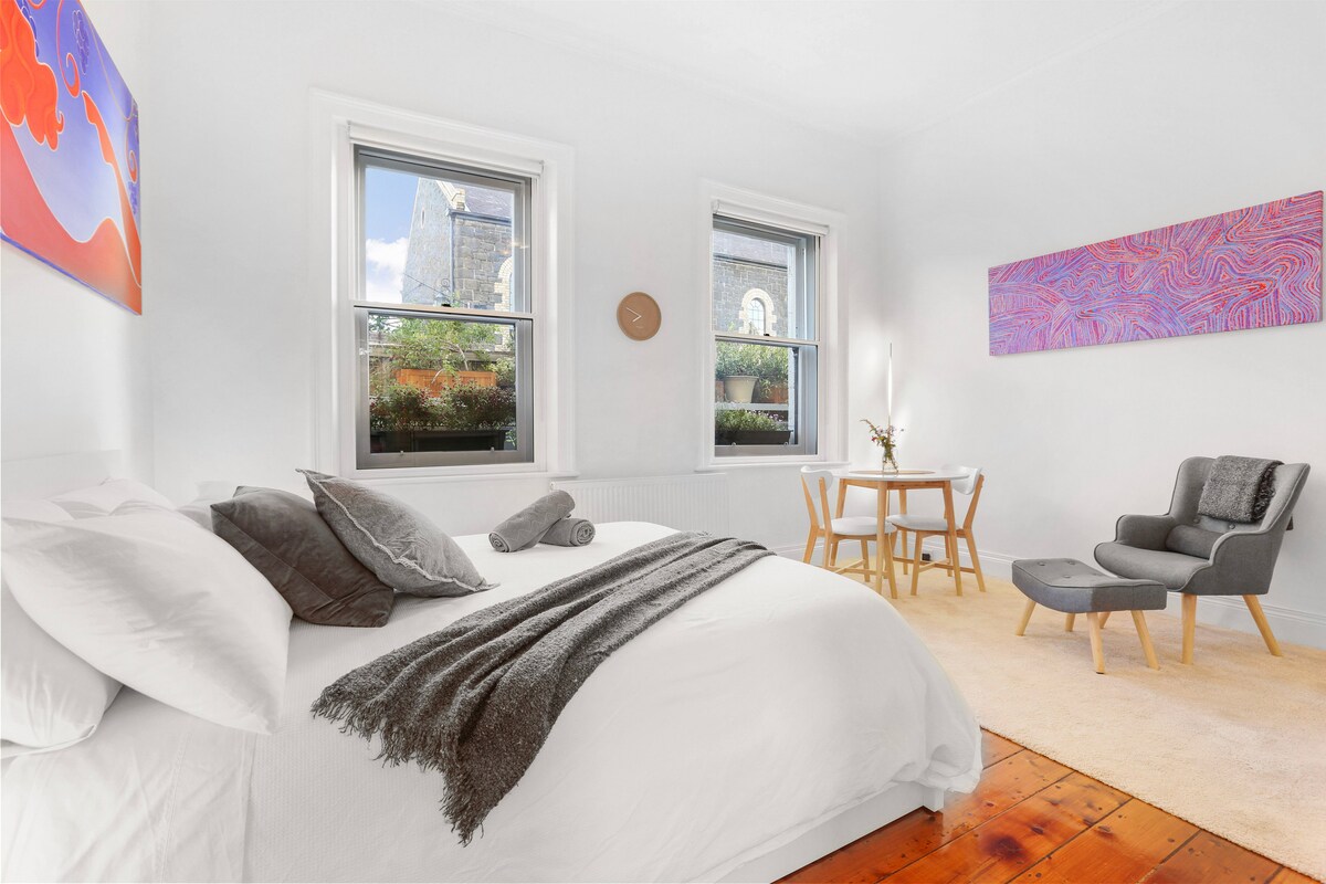 A bright room features a spacious bed adorned with crisp white linens and several plush pillows. Two windows provide natural light and views of the outdoors. An armchair and a small dining table are positioned nearby on a light carpet, complemented by colorful wall art.