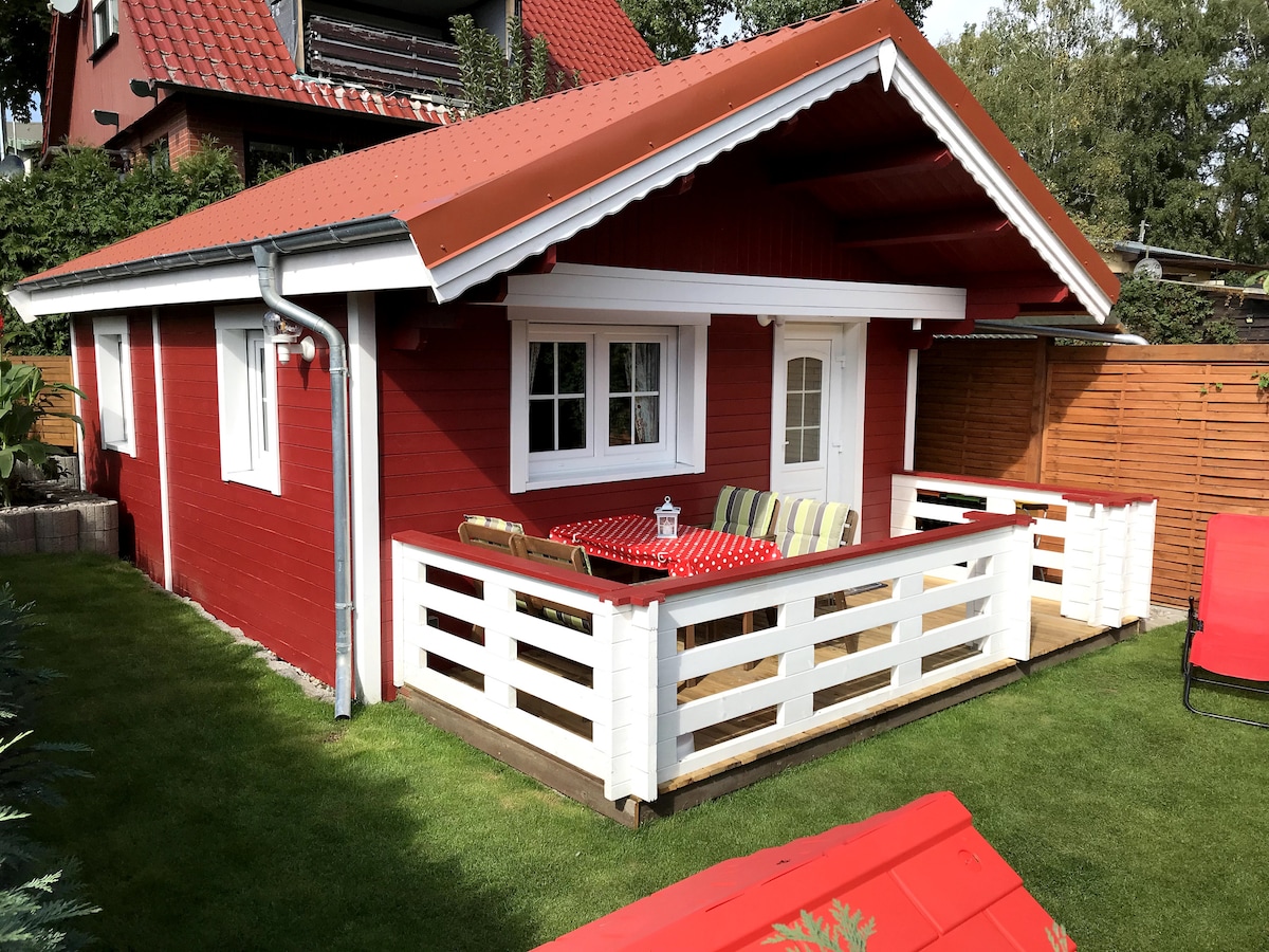 A charming red wooden house is surrounded by a grassy area and features a covered entrance with white railings. A table with colorful seating is positioned on the porch, inviting outdoor relaxation. Large windows allow natural light into the living space.