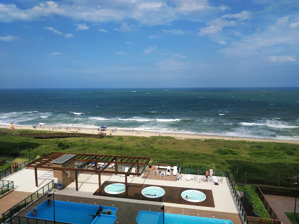 An expansive view of the coastline is showcased, with rolling waves gently lapping against the shore. In the foreground, several pools are visible on a landscaped deck, surrounded by lounge chairs and shaded areas. The horizon meets a clear blue sky dotted with a few clouds.