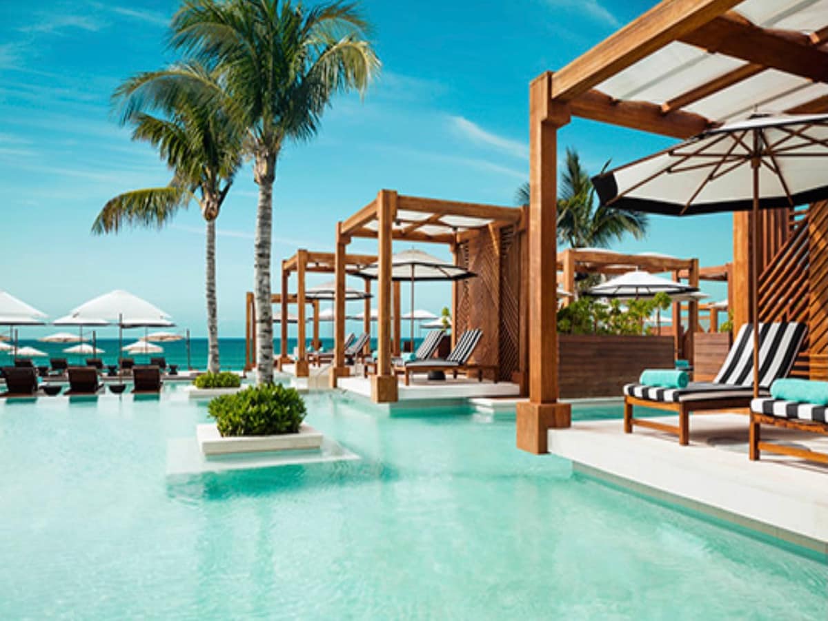 A tranquil pool area is surrounded by swaying palm trees and stylish lounge chairs. Canopy structures provide shaded seating, while large umbrellas are positioned over additional chairs. The clear blue water of the pool reflects the bright sky, creating a serene atmosphere for relaxation.
