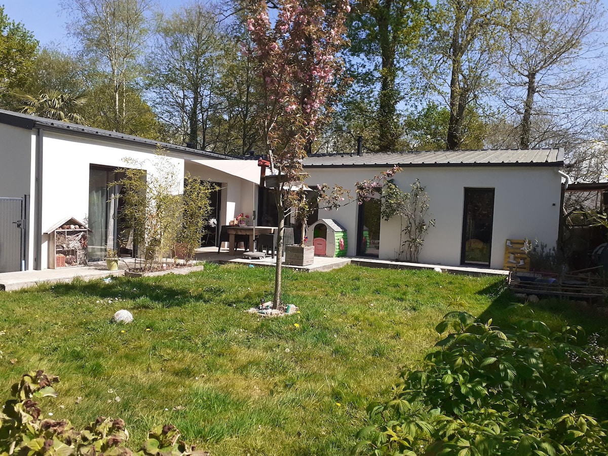 A spacious garden features a blend of grass and paved areas, framed by trees. Two modern structures are visible, with large windows allowing natural light inside. A picnic table and a bird feeder are positioned in the yard, adding functional elements to the outdoor space.