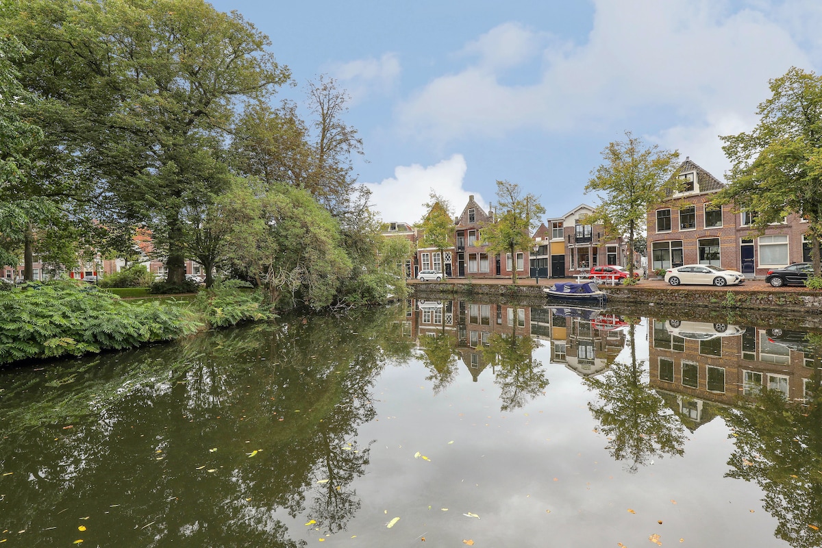 A serene canal scene reflects charming houses and lush greenery along its banks. Trees frame the water, creating a peaceful atmosphere. A few boats are moored nearby, contributing to the tranquil setting of this beautiful area.