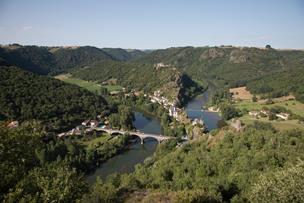 A panoramic view captures the peaceful village of Ambialet nestled along the Tarn River, surrounded by lush green hills. A bridge spans across the river, connecting both sides. The serene landscape features a blend of forests and open fields under a clear blue sky.