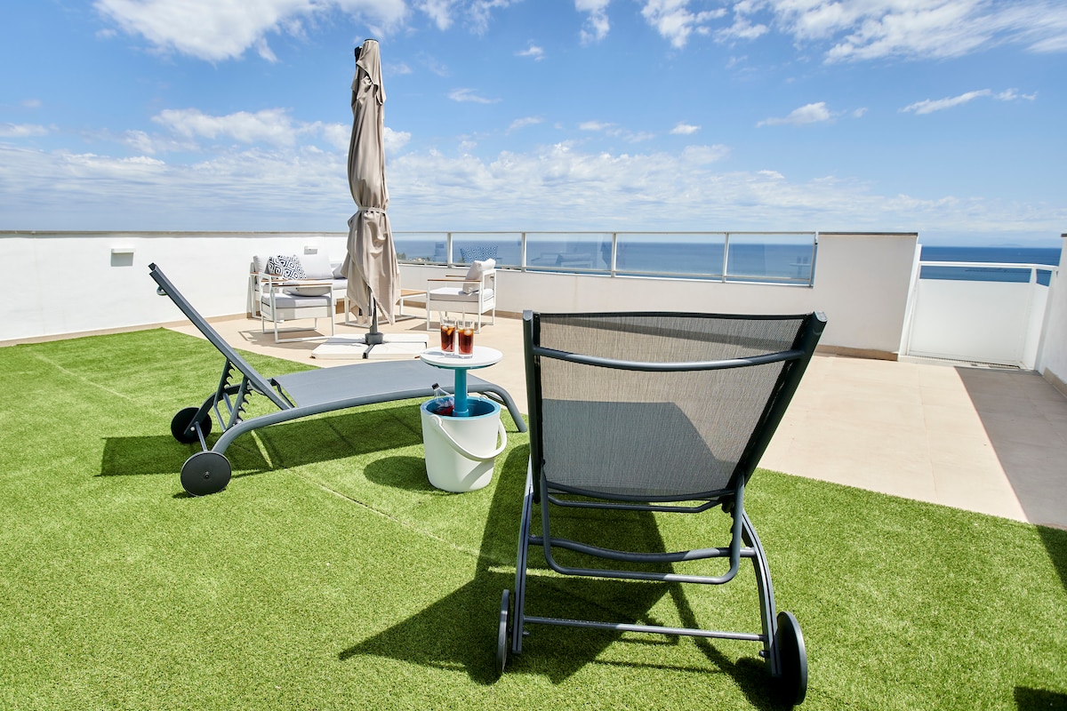 A spacious rooftop terrace is depicted, featuring comfortable lounge chairs and a small side table. The terrace is adorned with artificial grass, enhancing the inviting outdoor space. A large umbrella provides shade while panoramic views of the coastline are visible in the background.