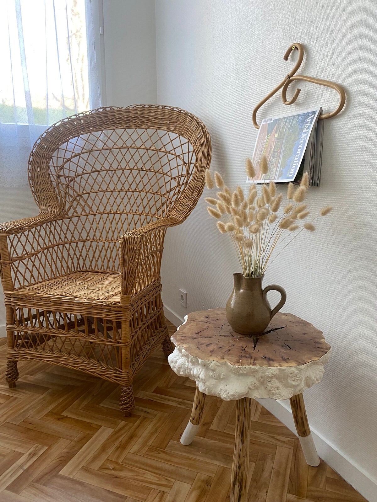A woven chair is positioned beside a small, unique table featuring a natural wood finish. A vase with dried flowers rests on the table, while a wall-mounted hanger holds a magazine, creating a cozy reading nook.