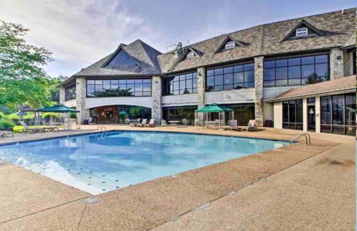 An outdoor pool is centered within a spacious patio, surrounded by lounge chairs and green umbrellas. The multi-story building features large windows that reflect natural light while providing views of the surrounding landscape.