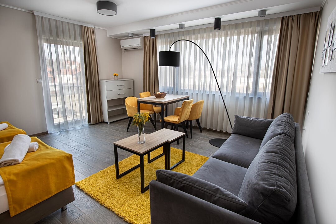 A spacious living area features a dark gray sofa paired with a coffee table on a soft yellow rug. A dining table with four yellow chairs is illuminated by a modern arc floor lamp. Large windows with sheer curtains allow natural light to fill the room.