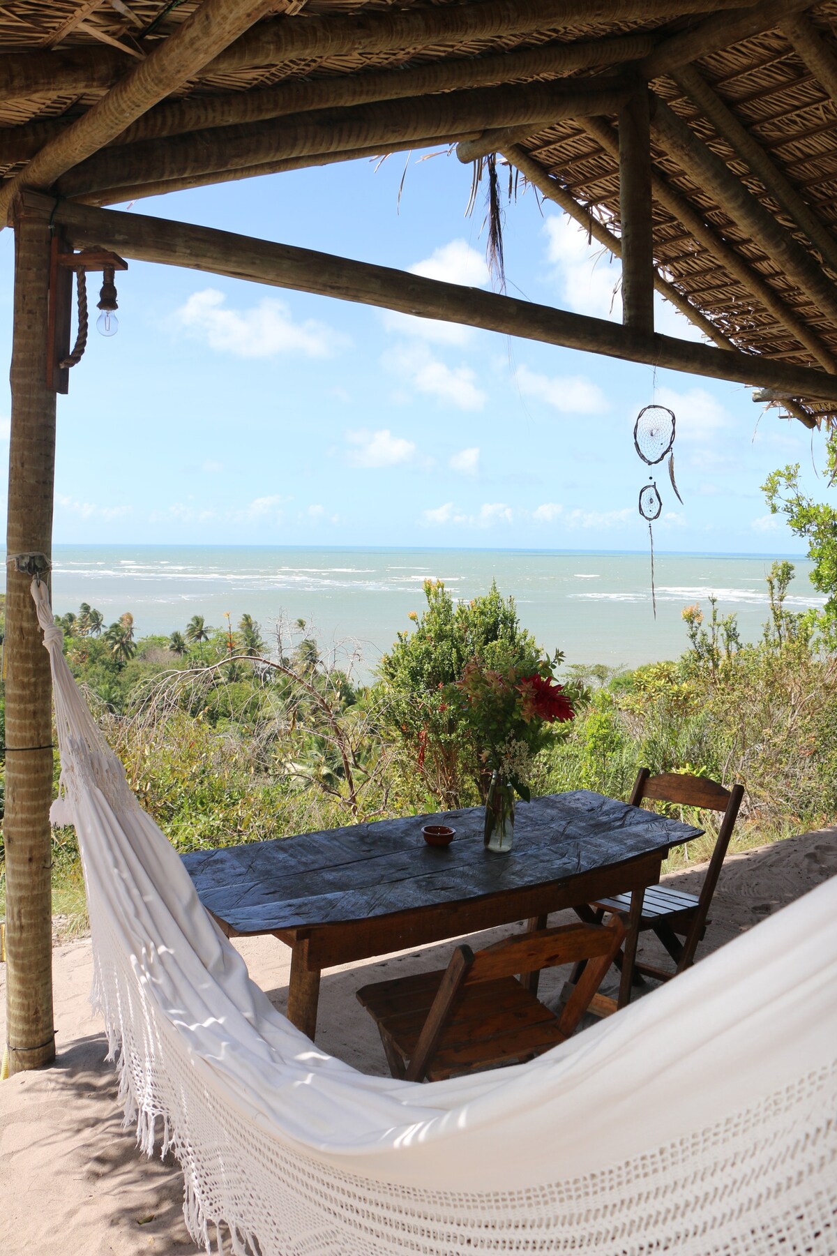 A shaded outdoor area features a wooden table accompanied by two chairs, positioned beneath a thatched roof. A hammock is draped nearby, with a vase of flowers adding a natural touch. The backdrop reveals a coastal view with greenery and distant ocean waves.