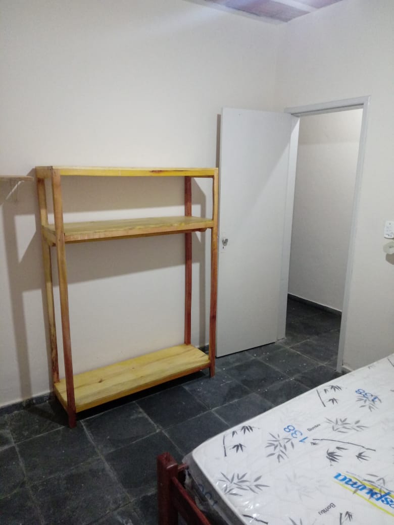 A simple room features a wooden shelving unit against one wall, providing open storage space. A bed with a floral-patterned mattress occupies the floor, while a door leads to another area of the unit. Neutral-toned walls and a tile floor add to the minimalist design.