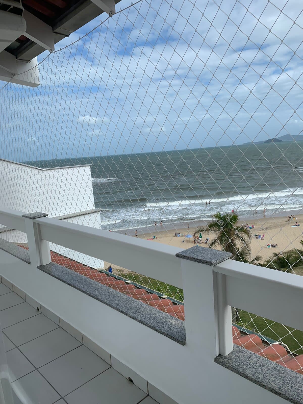 A balcony view reveals a sandy beach with gentle waves lapping at the shore. Palm trees are visible nearby, complementing the coastal scenery. The sky displays a mix of clouds and sunlight, offering a serene atmosphere. A netted barrier ensures safety while enjoying the view.