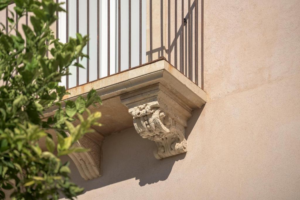 The image captures a detailed architectural feature of a corner balcony, showcasing a sculpted stone bracket that supports the balcony above. A bright green leafy branch is visible in the foreground, providing a natural contrast to the soft beige façade of the building.