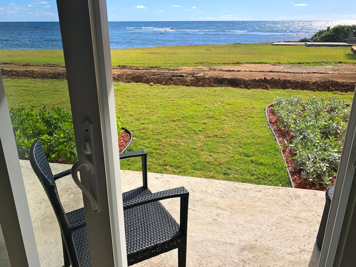 A view from an open doorway reveals a well-maintained grassy area leading to the ocean. Two outdoor chairs are positioned on a concrete patio, offering a serene spot to enjoy the surrounding landscape and ocean horizon.