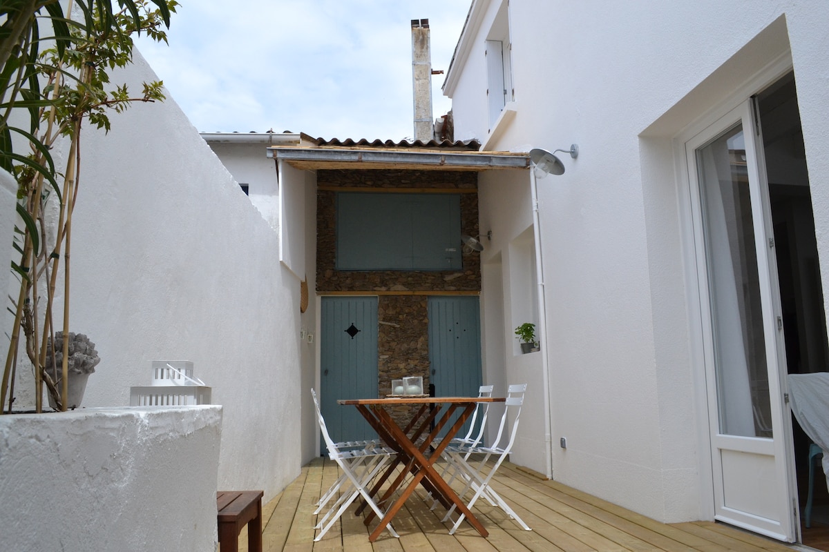 A wooden terrace is situated between white-walled structures, featuring a rectangular table surrounded by six white chairs. A rustic stone accent wall is visible in the background, adding texture to the space. Soft natural light illuminates the area, creating a serene atmosphere.