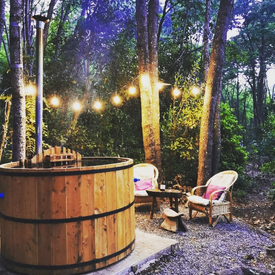 A wooden hot tub surrounded by trees and soft lighting creates a cozy outdoor space. Two wicker chairs are positioned beside a small table, enhancing the serene atmosphere of the forest setting.