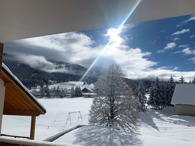 A winter landscape is revealed from the terrace, showcasing snow-covered mountains and trees under a partly cloudy sky. Sunlight glimmers on the snow, providing a serene view of the surrounding countryside, including distant homes and a playground swing.
