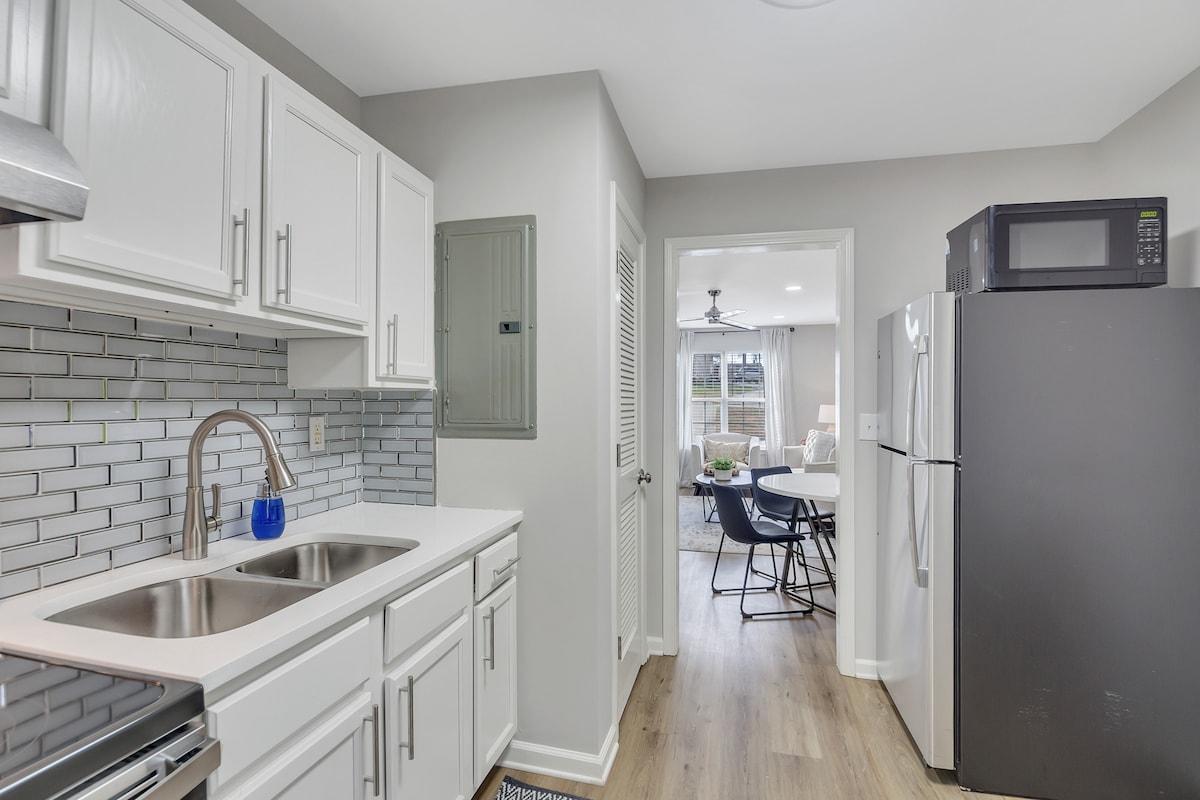 A clean kitchen features white cabinetry and stainless steel appliances. A classic tile backsplash complements the modern fixtures. The adjacent hallway opens to a bright living area, providing easy flow between spaces.