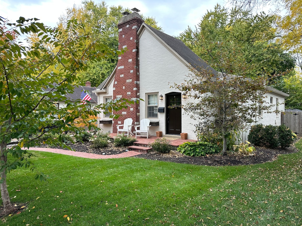 The exterior of the bungalow features a charming combination of brick and white siding, framed by lush greenery. A landscaped front yard includes a path leading to the entrance, with two white chairs positioned invitingly on the porch.