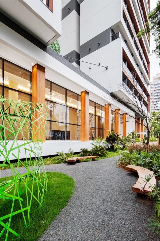 An outdoor pathway meanders through green landscaping, bordered by a mix of lush plants and benches. A modern, geometrically designed green sculpture adds a unique touch near the building's expansive glass windows, which reflect the surrounding environment.