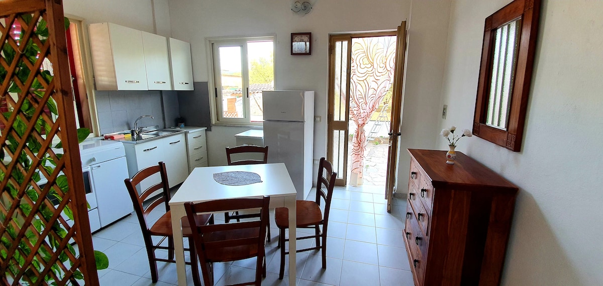 An open kitchen and dining area is presented, featuring a white table with four wooden chairs. Natural light enters through a window and a door leading to an outdoor space. A refrigerator and an oven are visible, along with wooden cabinets enhancing functionality.