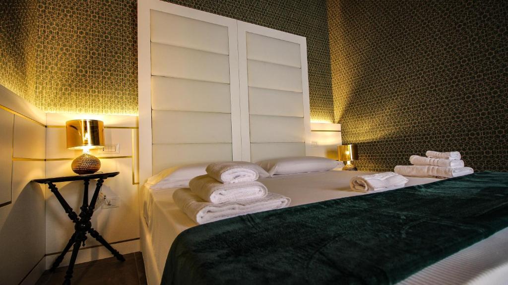 A spacious bed with a muted green bedspread is flanked by two modern bedside lamps, casting a soft glow. A stack of neatly arranged towels sits on the edge of the bed, while a patterned wallpaper adds a touch of elegance to the room.