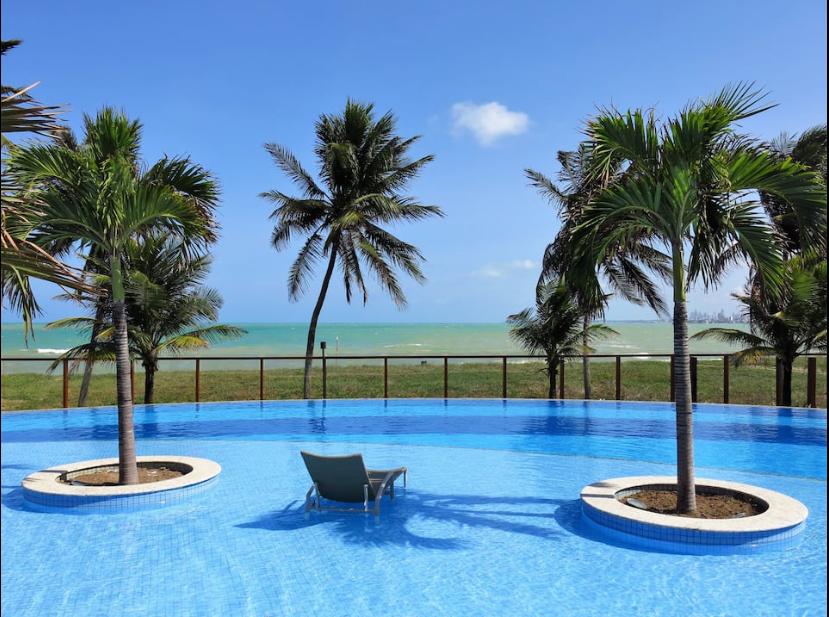 A serene swimming pool is surrounded by palm trees, offering a view of the ocean beyond. The water's surface reflects the clear blue sky, creating a calming atmosphere. A single lounge chair rests on the pool's edge, inviting relaxation.