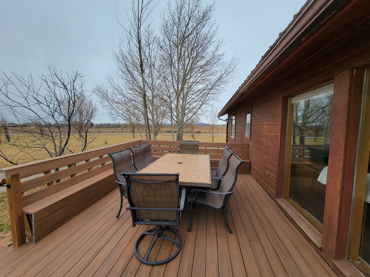 A wooden deck features a large dining table surrounded by comfortable chairs. The view beyond includes open pasture and distant trees, creating a relaxed outdoor dining space. Nearby, the structure's exterior showcases warm wood tones and large glass windows, enhancing the connection to the surrounding landscape.