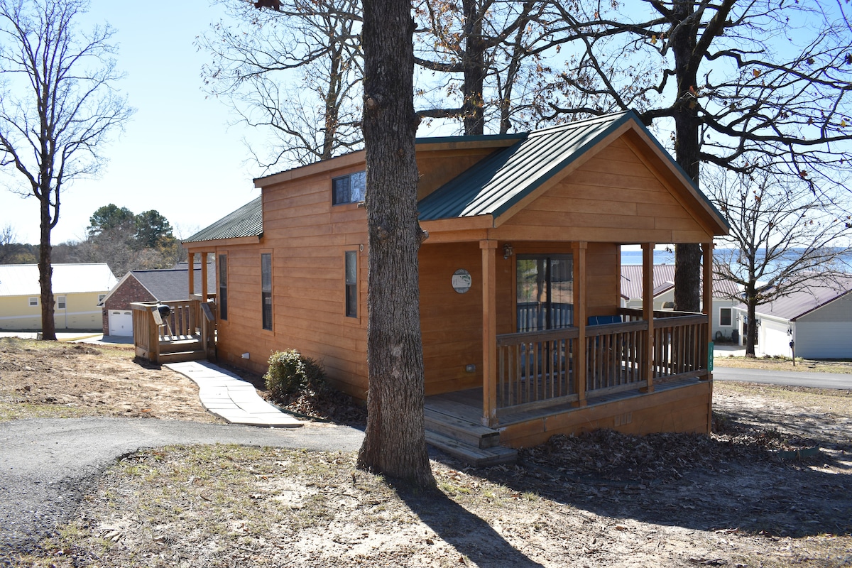 The cabin is constructed with warm wood siding and features a green metal roof. A spacious porch is visible, inviting relaxation. The surrounding area includes trees and a pathway leading to the entrance, while neighboring homes can be seen in the distance.
