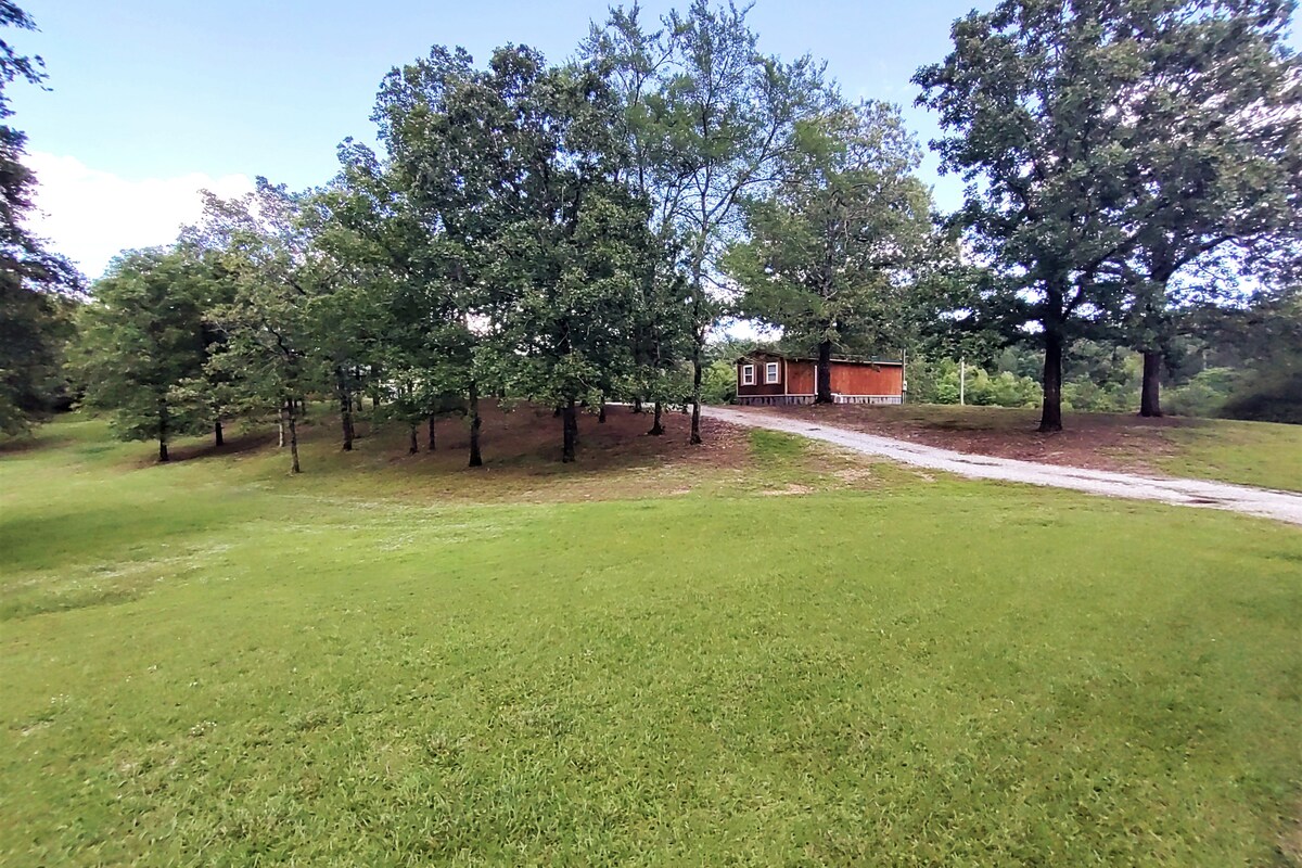 A spacious lawn area features well-maintained grass and several trees providing shade. The pathway leads to the guest house, which is set back among the trees. The surrounding landscape consists of gentle hills, creating a serene and natural environment.