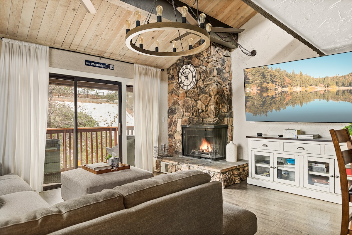 A cozy living area is designed with a stone fireplace as a focal point. Modern furnishings include a sectional sofa and a coffee table. Large windows provide natural light and views of the snowy landscape. A wall-mounted TV is positioned above a white media console.
