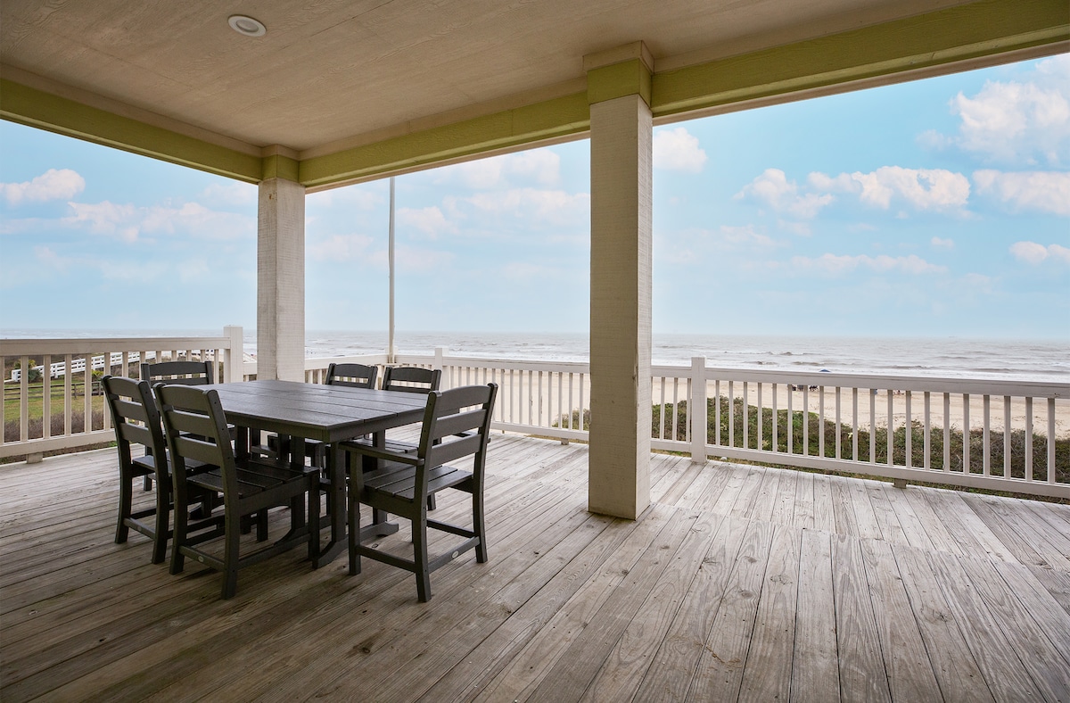A spacious outdoor deck is showcased, featuring a large wooden dining table surrounded by six chairs. The ocean view is visible beyond the railing, complemented by a bright sky with soft clouds, creating a serene atmosphere for outdoor dining.