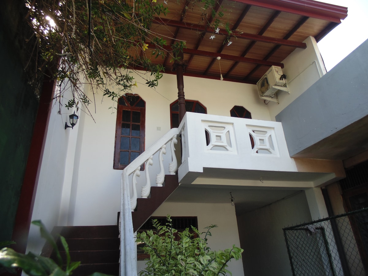 An outdoor view of a two-story structure showcases a staircase leading to an upper level with decorative white railings. Windows with arched tops provide natural light, while greenery surrounds the base of the stairs, creating a tranquil atmosphere.
