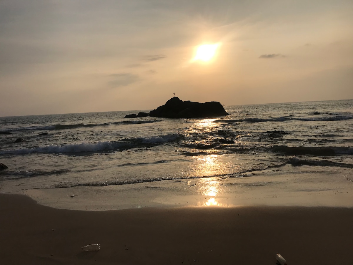 A serene beach scene is depicted with gentle waves lapping against the shore. A solitary rock is visible in the water, partially illuminated by the soft glow of the setting sun. The sky showcases a gradient of warm colors, reflecting tranquility.