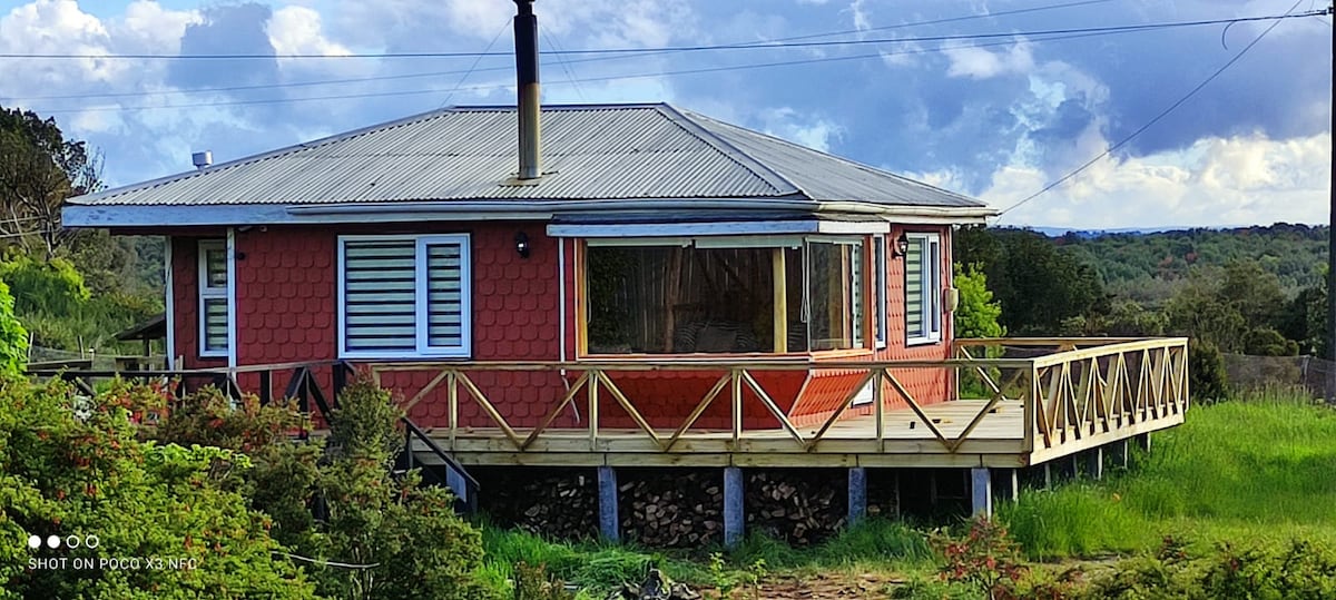 The cabin is situated on a raised structure, surrounded by lush greenery. A spacious wooden deck extends from the exterior, featuring a railing and providing ample outdoor space. Large windows allow for natural light, while a chimney rises from the roof.